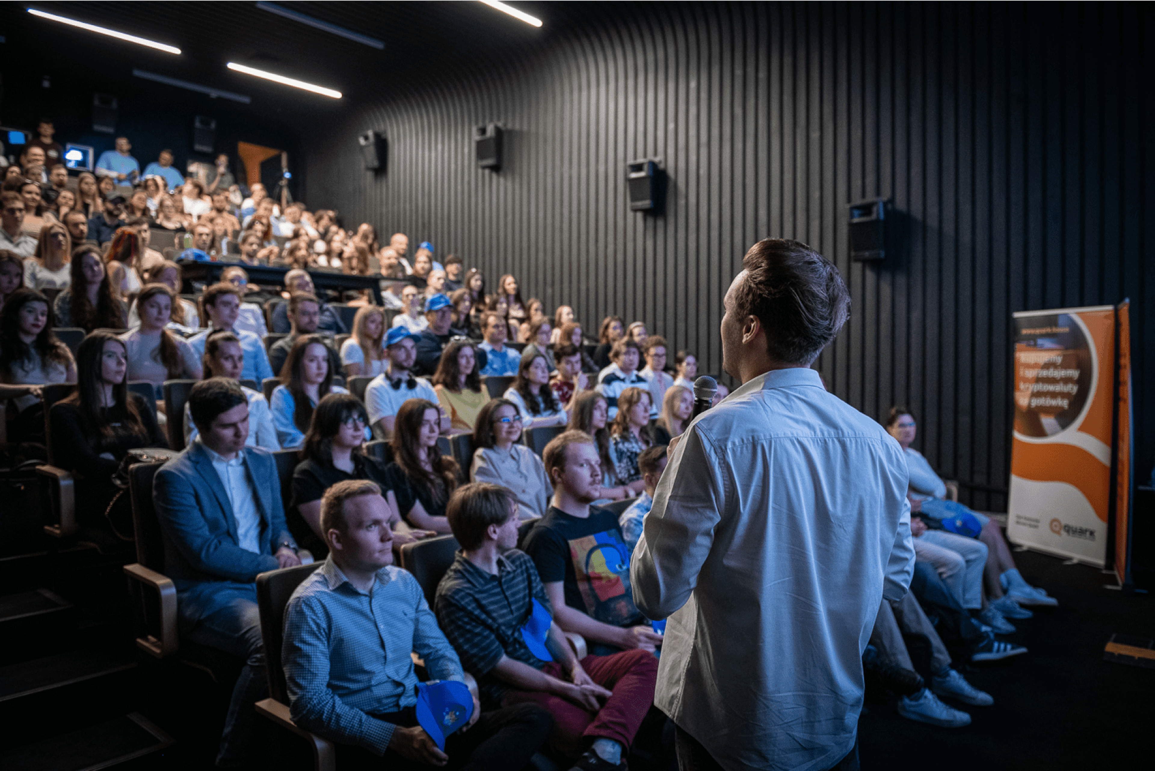 Speaker presenting to a full audience in a dark auditorium, with attendees attentively listening and engaged in the presentation.