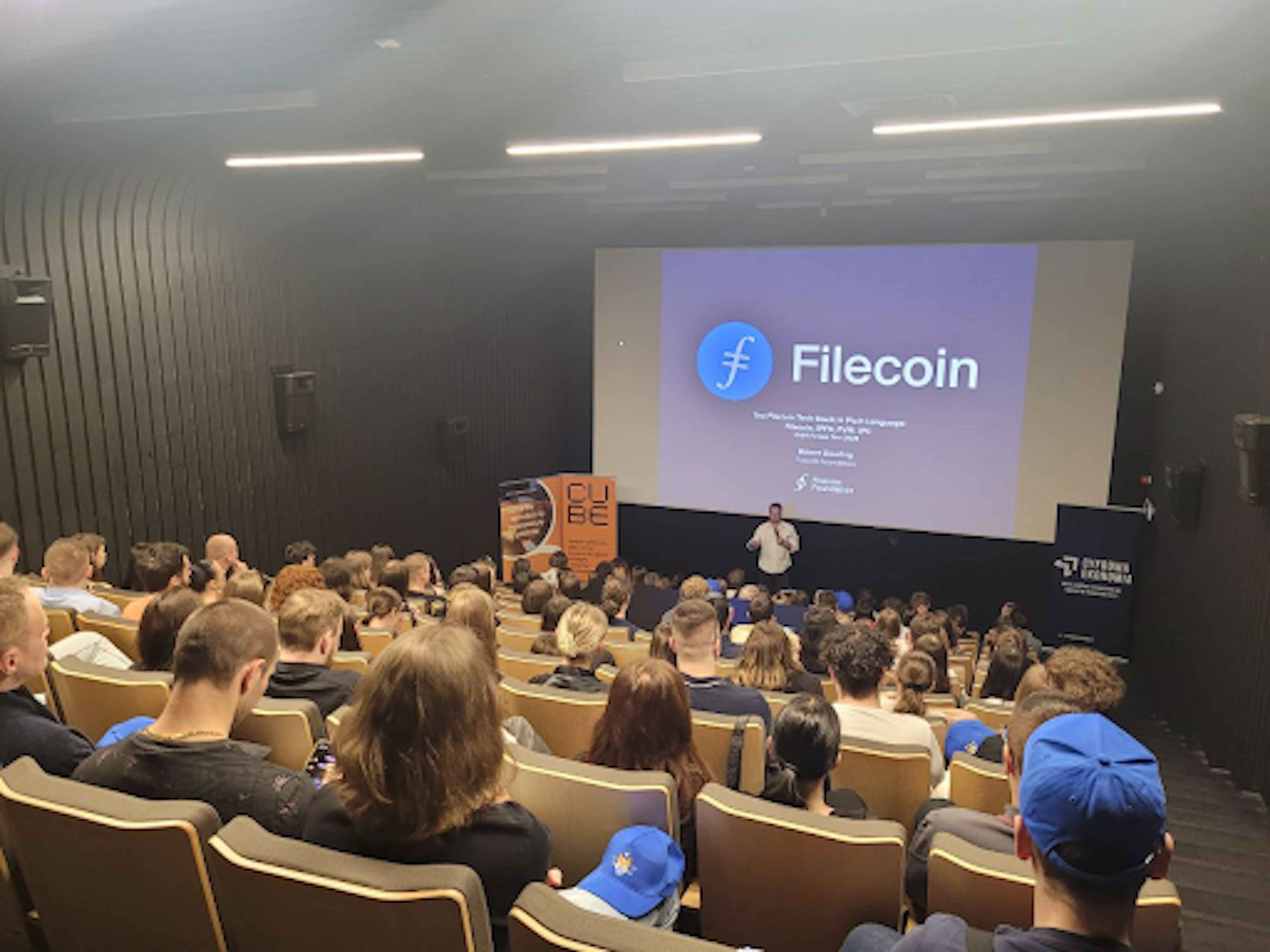 A person is giving a presentation on Filecoin to an audience in a lecture hall, with a slide displaying the Filecoin logo and title.